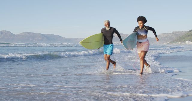 Senior couple enjoying a sunny day at the beach, carrying surfboards and running into ocean waves. Great for promoting active lifestyles, retirement activities, outdoor adventures, and fitness for older adults. Perfect for travel agencies, wellness blog posts, or advertisements targeting active seniors.