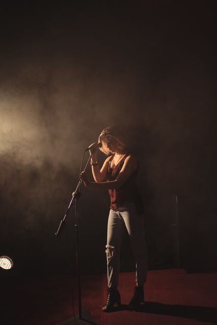 Confident female singer performing on stage with a microphone. She is wearing jeans and boots, standing under a spotlight with a smoky background. Ideal for use in music-related promotions, concert advertisements, entertainment blogs, and artist portfolios.