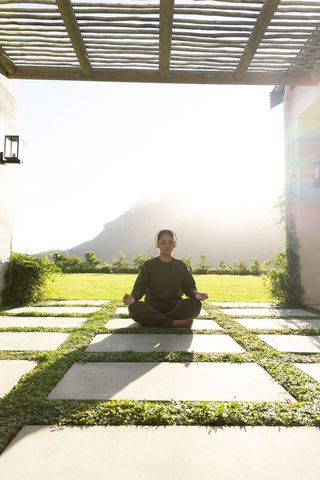 Asian woman meditating on sunlit patio under pergola wearing headphones and yoga wear