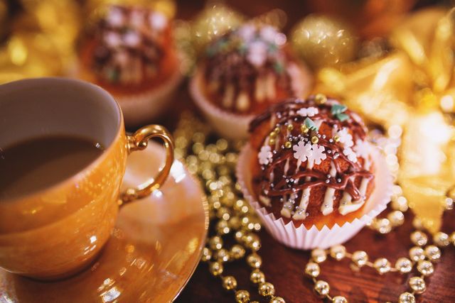 Close-up view of beautifully decorated cupcakes with chocolate drizzle and festive sprinkles, accompanied by a warm cup of coffee on a table adorned with gold beads and Christmas ornaments. Ideal for seasonal promotions, holiday recipes, Christmas-themed advertising, and festive greeting card designs.