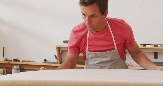 Middle-aged male carpenter engaged in shaping wooden surface in a workshop setting. Wearing a pink t-shirt and apron, demonstrating focused craftsmanship with tools and workbench in the background. Useful for illustrating skilled manual labor, woodworking process, and professional craftsmanship.