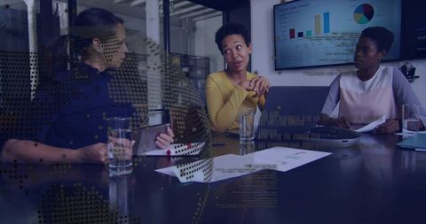 Woman in yellow sweater gesturing while team reviewing sales charts on laptop and monitor
