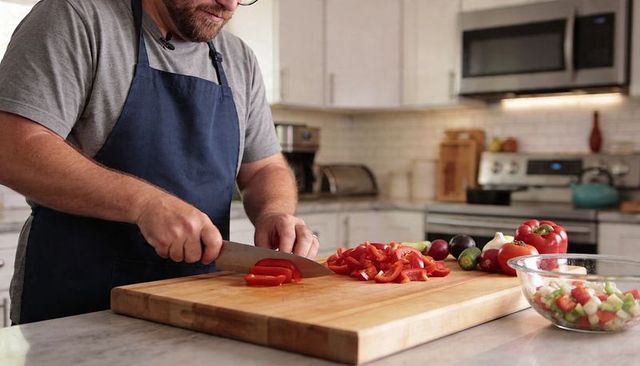 Bearded man wearing apron and glasses chopping red bell pepper on wooden cutting board
