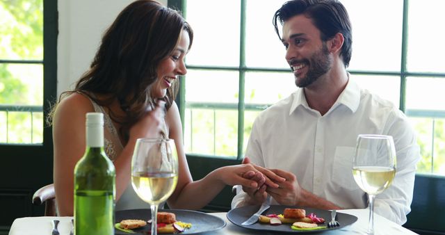 A young Caucasian couple enjoys a romantic meal together in a bright dining setting, with copy space. They are sharing a moment of affection, holding hands across the table with smiles and a bottle of wine enhancing the intimate atmosphere.