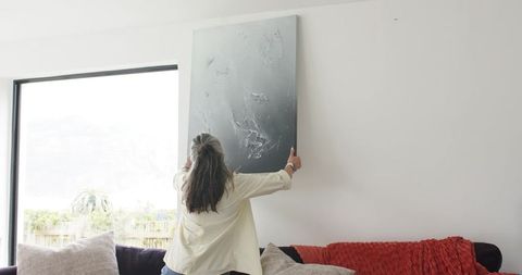 Mature woman hanging abstract dark canvas above sofa in bright contemporary living room