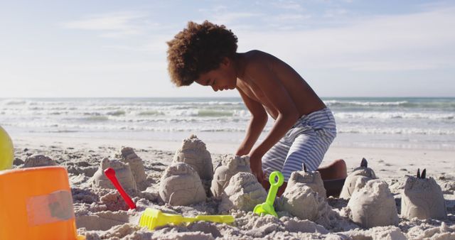 Young boy enjoying a sunny day at the beach, building intricate sandcastles near the shoreline. Ideal for content related to summer vacations, child play, beach activities, family outings, and outdoor activities.