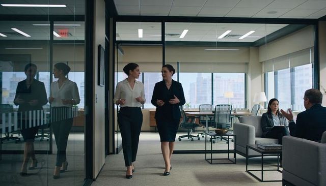 Two professional women walking and chatting in modern glass-walled office corridor