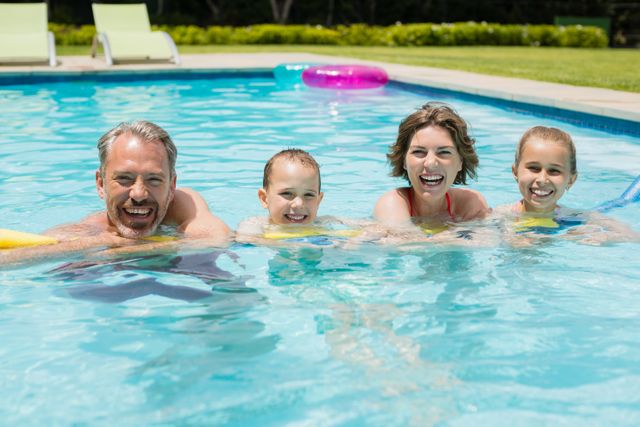 This image shows a happy family of four enjoying swimming in an outdoor pool on a sunny day. The parents and two children are smiling and appear to be having fun. This image can be used for promoting summer activities, family vacations, outdoor events, or swimming pool advertisements.