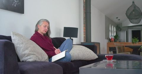 Senior woman writing notebook on sofa, cozy living room, red tea, modern interior