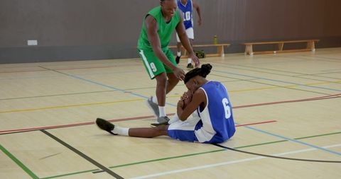 Basketball player holding knee following fall while teammate helping on indoor court