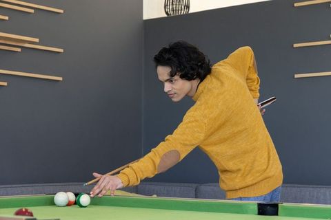 Young man leaning over pool table aiming cue while holding phone in mustard sweater