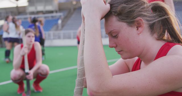 Female field hockey players experiencing disappointment after losing a game. One player with head down, clinging to her stick, while another sits down in the background. Could be used to illustrate themes of sports psychology, teamwork, determination in the face of setbacks, or the emotional impact of losing in competitive sports.