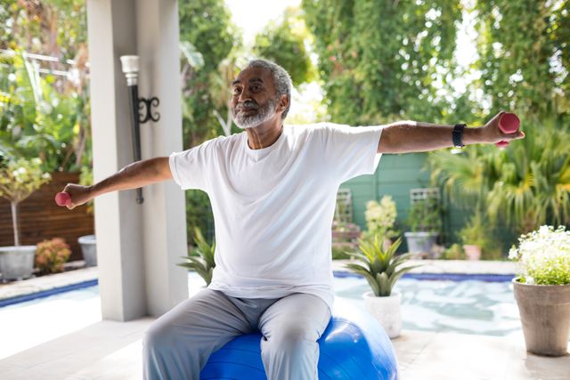 Senior man lifting dumbbells while sitting on a stability ball in a yard. Ideal for promoting senior fitness, healthy lifestyle, and outdoor exercise. Useful for fitness blogs, health and wellness articles, and advertisements targeting active aging and home workout routines.