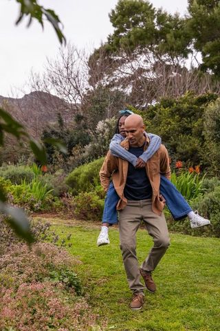 African american man carrying child piggyback walking through lush garden park