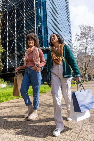 Urban friends walking on sunny sidewalk with shopping bags by modern glass office tower