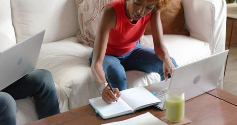 Focused woman writing in notebook while working on laptop in cozy living room workspace