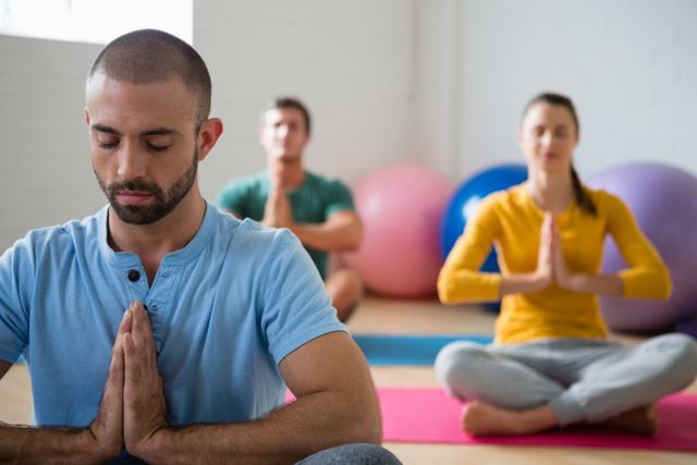 Yoga instructor leading a meditation session with students in a health club. Participants are sitting on mats in a prayer position, focusing on mindfulness and relaxation. Ideal for use in content related to fitness, wellness, mental health, and lifestyle.
