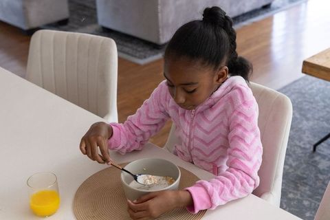 Young african american girl eating cereal at dining table in pink hoodie morning breakfast