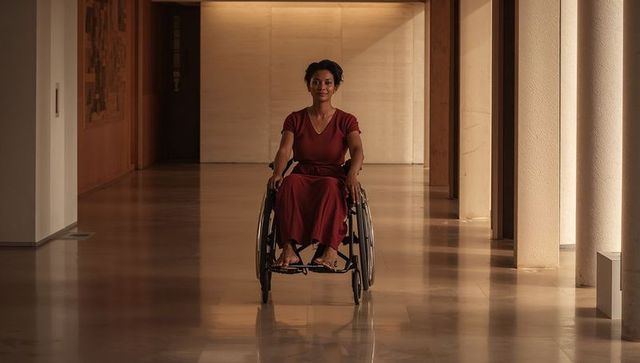 Woman pushing wheelchair down sunlit modern corridor wearing maroon dress