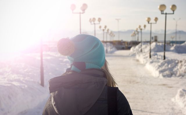 Person walking outside on a sunny winter day. Snow covers the ground and white lamp posts line a paved pathway. The person is wearing a blue beanie and warm winter clothing. This can be used for promoting winter tourism, outdoor activities, or capturing the beauty of winter walks.