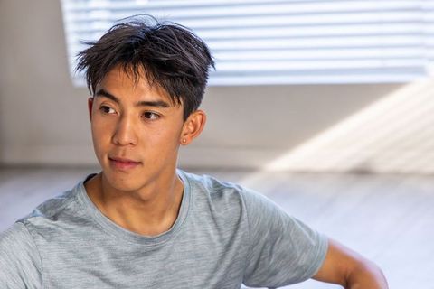 Young asian man sitting in sunlit room looking sideways wearing casual grey tee