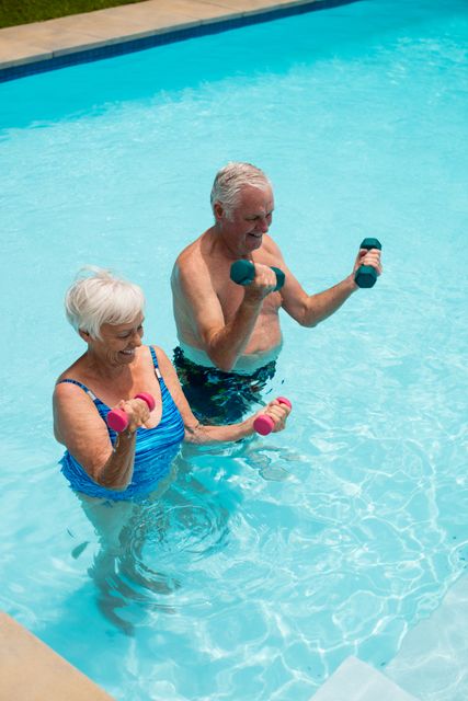 Senior couple enjoying a fitness routine with dumbbells in a swimming pool. Ideal for promoting active lifestyles, senior wellness programs, retirement community advertisements, and health-related articles focusing on the elderly staying fit and happy. The cheerful expressions and bright setting suggest health and vitality.