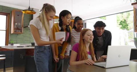 Diverse teens gathering around laptop at sunlit kitchen island sharing study and laughter