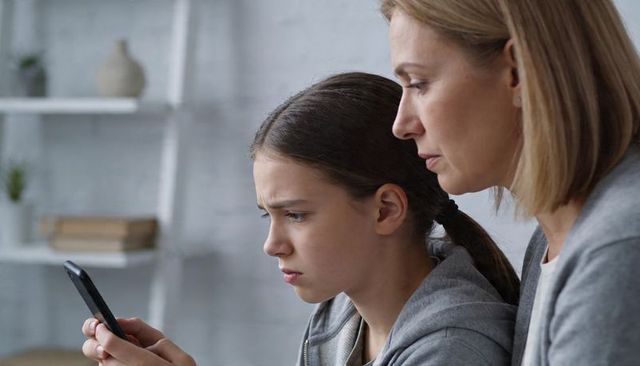 Concerned mother guiding worried teen holding smartphone and frowning in cozy home