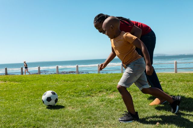 Father and son enjoying a sunny day playing soccer on a grassy area near the beach. Perfect for illustrating family bonding, outdoor activities, and summer fun. Ideal for use in advertisements, family lifestyle blogs, and promotional materials for outdoor sports and recreation.