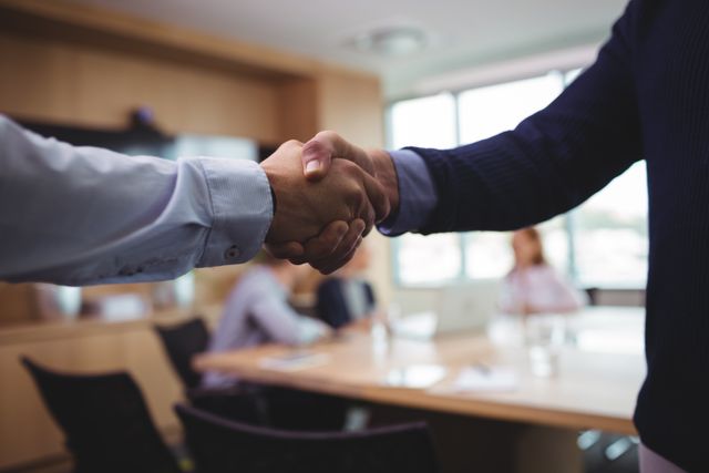 This image shows a close-up of two business people shaking hands in a board room, symbolizing agreement, partnership, and successful negotiations. The background features a blurred view of a meeting with other professionals, indicating a collaborative and professional environment. This image can be used for business-related content, articles on corporate culture, teamwork, partnership announcements, and promotional materials for business services.