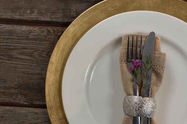 Close-up of an elegant table setting featuring silverware wrapped in a beige napkin with a small purple flower, placed on a white plate with a gold charger. The wooden plank background adds a rustic touch. Ideal for use in articles or advertisements related to dining, hospitality, event planning, or home decor.