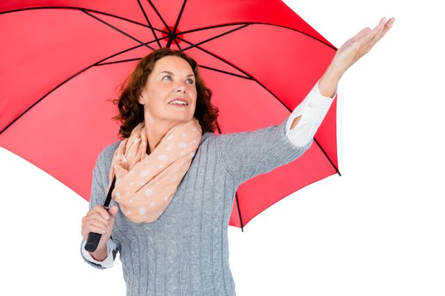 Mature woman holding a red umbrella and looking up with a smile. She is wearing a grey sweater and a light pink scarf, standing against a white background. This image can be used for weather-related content, advertisements for rain gear, or articles about staying positive during rainy days.