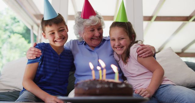 Caucasian senior woman in party hat hugging her grandchildren with birthday cake on table at home. social distancing during coronavirus quarantine lockdown.
