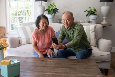 Senior chinese couple sharing smartphone and smiling sitting on cozy living room sofa