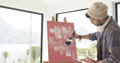 Indian man painting abstract white brushstrokes on red canvas in bright lakeside studio