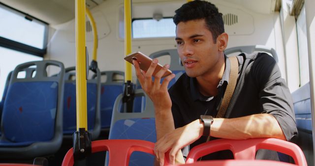 Young man in black shirt riding public bus, holding smartphone close to his face, and talking. Open seats and urban setting in background. Ideal for illustrating public transportation, commuting lifestyles, use of technology on-the-go, and modern urban life.