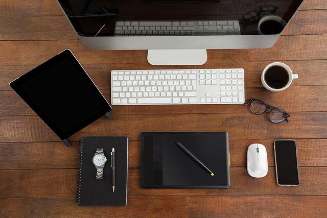 Overhead view of a modern office desk featuring a computer, digital tablet, smartphone, keyboard, mouse, coffee cup, glasses, watch, notebook, and stylus. Ideal for illustrating concepts related to technology, business, productivity, remote work, and modern workspaces.