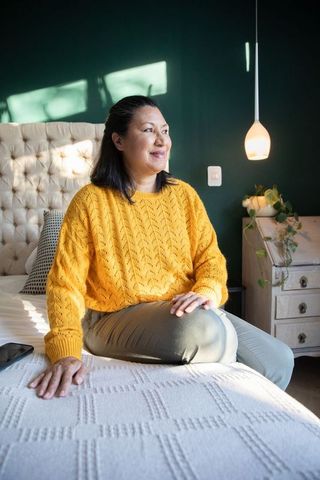 Serene middle-aged woman wearing mustard sweater sitting on bed facing sunlit window