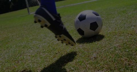 Closeup soccer player kicking ball with black cleats and blue socks on sunlit grass near goal