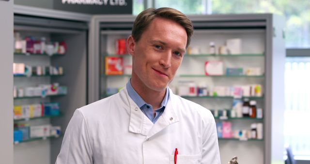 Male pharmacist wearing white coat standing and smiling at pharmacy counter, shelves with medication in background. Ideal for use in advertisements, educational materials, healthcare websites, and articles about pharmaceutical services, customer service, and medication management.