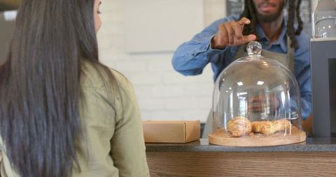Barista reaching for croissants under glass cloche while customer waits at cafe counter