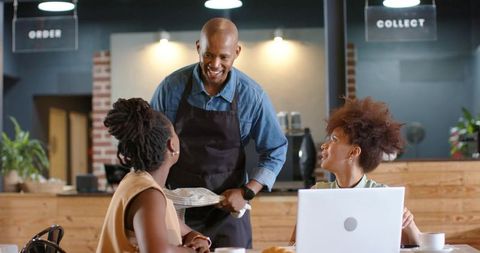 Smiling barista leaning toward patrons in cozy cafe with laptop, cups, friendly service
