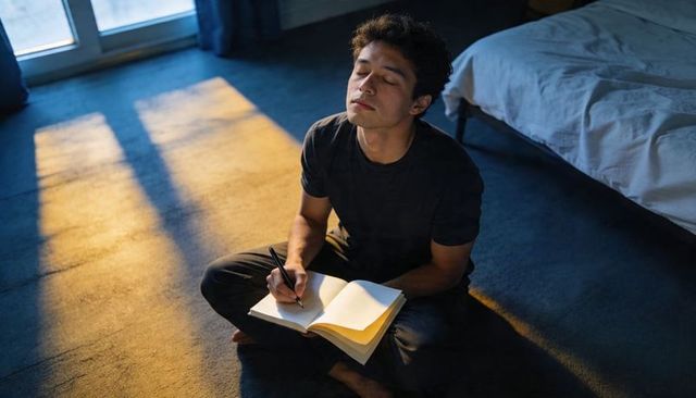 Young man journaling on bedroom floor at sunrise, warm sunlight and calm reflection
