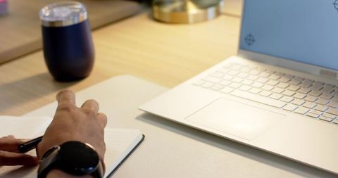 African american man writing notes at home desk with laptop keyboard, tumbler, warm light
