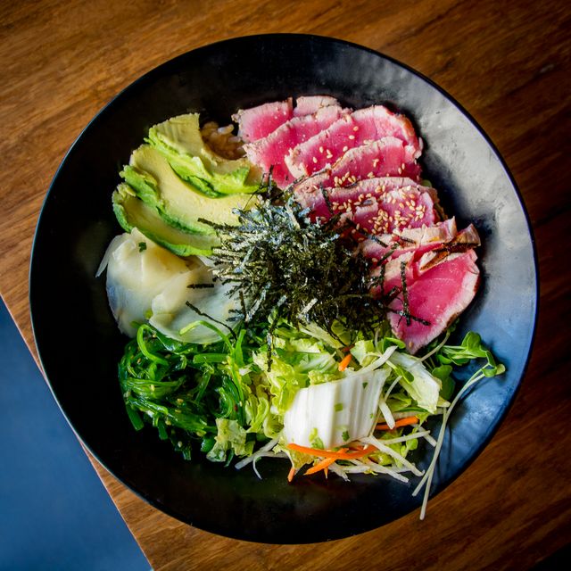 A vibrant ahi tuna poke bowl features fresh slices of ahi tuna and avocado arranged on a bed of rice, highlighted by mixed vegetables and seaweed. Light sesame seed garnish adds texture and visual appeal. Suitable for food blogs, healthy eating articles, Japanese cuisine demonstrations, and menu designs.