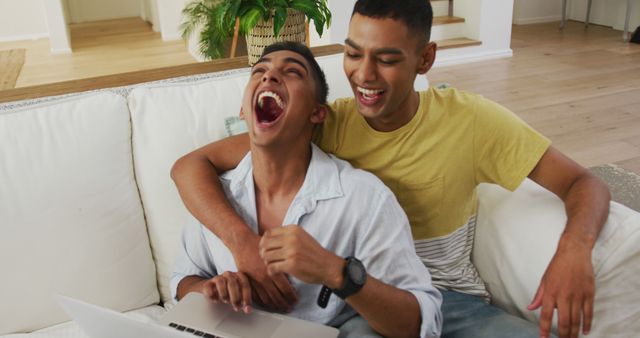 Smiling biracial gay male couple sitting on sofa using laptop and laughing. staying at home in isolation during quarantine lockdown.