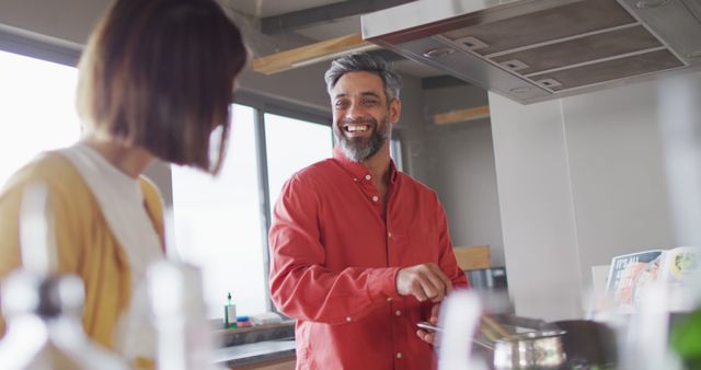 Middle-aged man cooking and smiling while engaging in conversation in a contemporary kitchen. Wearing a casual red shirt and sharing a moment of joy and relaxed lifestyle. Ideal for use in home life, joyful moments, culinary experiences, and positive lifestyle promotions.