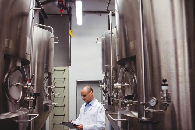 Brewery worker in white coat inspecting large stainless steel storage tanks while writing on clipboard. Ideal for illustrating industrial processes, quality control in brewing, and professional manufacturing environments.