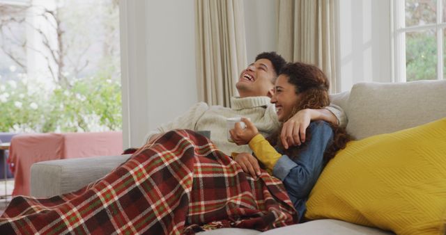 A couple is sitting on a couch, sharing a blanket and laughing together in a bright and cozy living room with large windows. This image can be used to depict themes of romance, comfort, home life, and warmth. Ideal for advertisements or articles about home decor, lifestyle, relationships, or cozy winter settings.