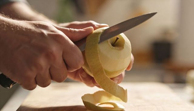 Hands peeling green apple into spiral with paring knife on wooden board